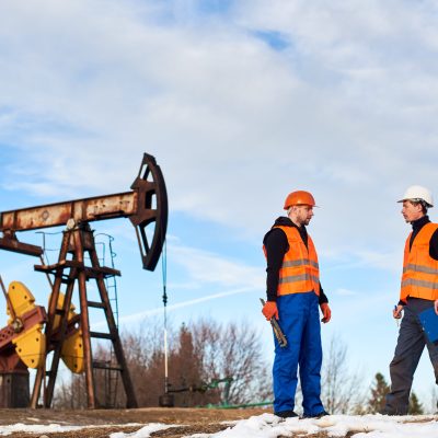 Two oil men in helmets and work vests standing near oil well pump jack and discussing work. Oil worker holding clipboard and talking with colleague at oil field. Concept of petroleum industry.